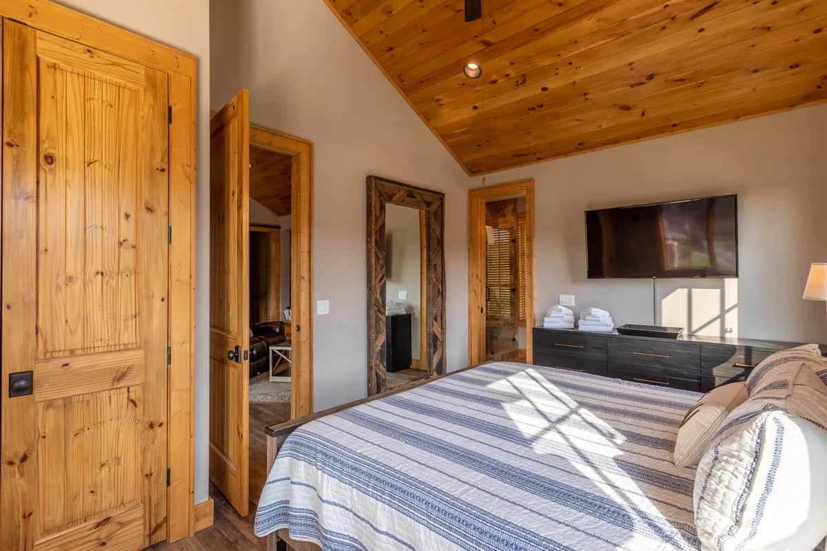 This image captures a serene bedroom scene featuring prominent wooden beams on the ceiling, a large bed with striped linens, and a mirror reflecting natural light throughout the space. The mood is calm and inviting, with hints of relaxation and comfort, ideal for both restful nights and cozy family gatherings.