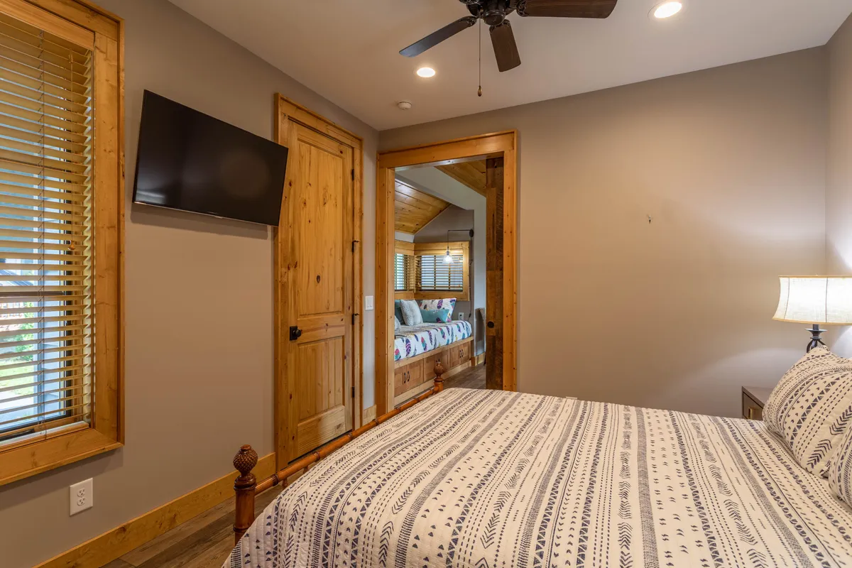 This image shows a cozy bedroom with a bed featuring patterned bedding, a mounted flat-screen TV, a wooden door, a window with blinds, and a warm lamp. The space feels inviting and is clearly designed for relaxation and restful nights after a day of adventure.