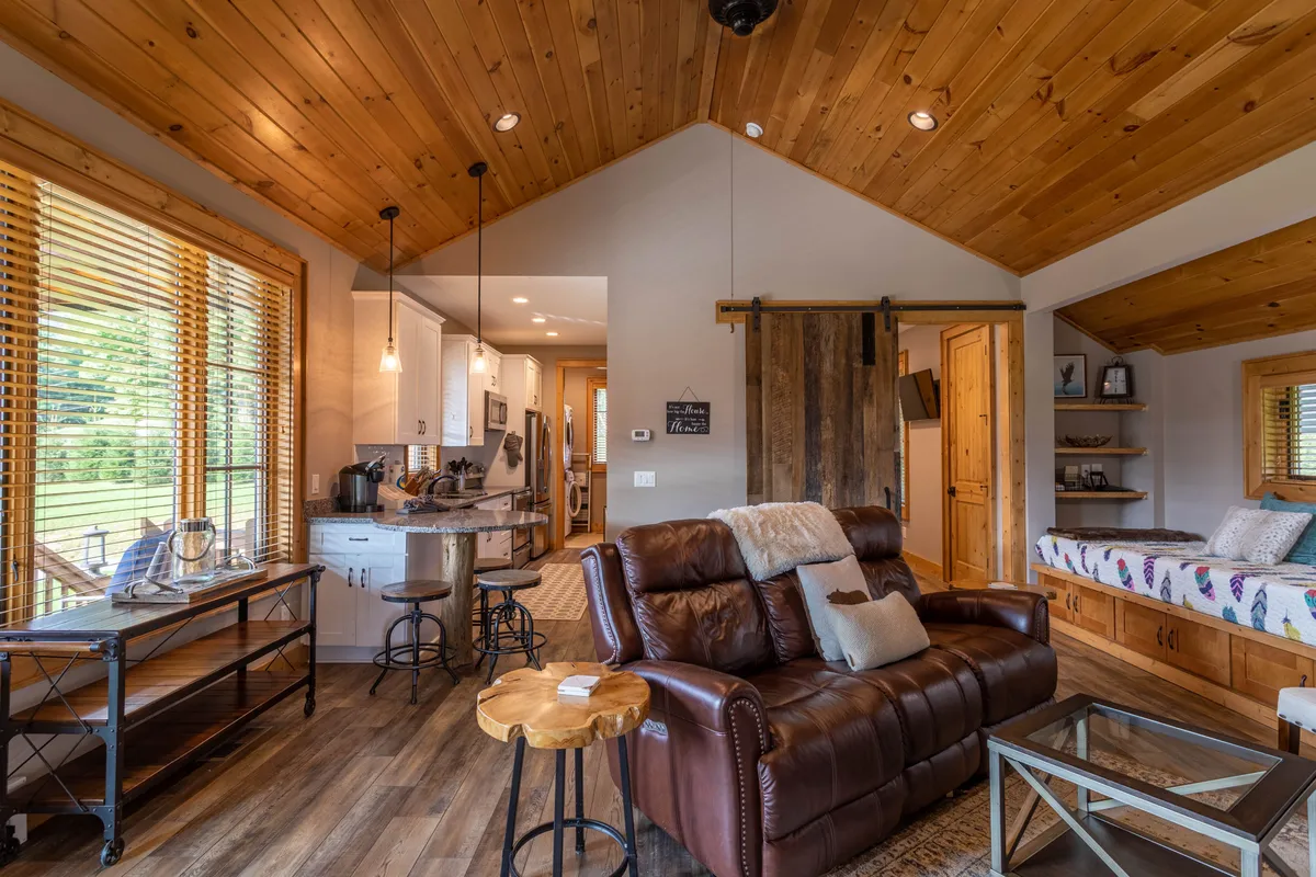 An inviting view of the living room with a luxurious leather sofa, wooden ceilings, large windows with scenic views, and a glimpse into the kitchen. The sofa is clearly visible, suggesting comfort and modern amenities for guests.