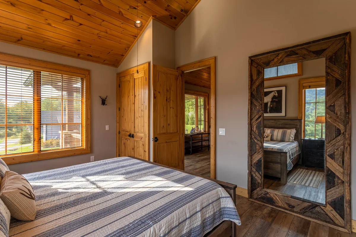 This interior scene features a comfortable bed with striped bedding, a large wooden mirror, and a sunlit window with blinds. The visible door and warm lighting create a welcoming space ideal for relaxation, reading, or quiet moments after a day exploring.