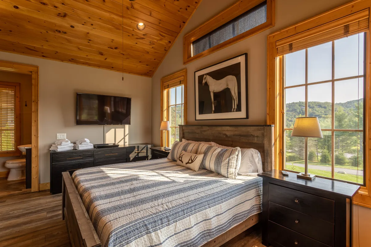 A spacious bedroom with a plush bed, large windows showcasing the outside light, tasteful artwork, and nightstands. The windows hint at the surrounding natural scenery, creating a tranquil retreat.