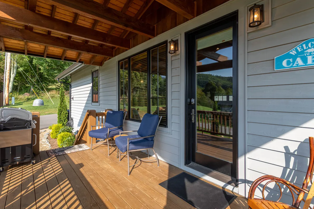 The inviting front porch features comfortable outdoor seating, a grill, and a welcoming sign. The porch is set against a backdrop of nature, suggesting a warm, friendly arrival and opportunities to enjoy meals or gatherings outside even in the winter season.